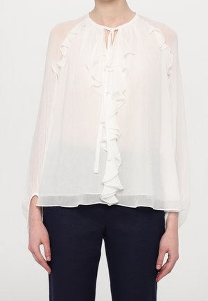 White sheer blouse with front ruffled trim and tie neckline, paired with dark navy pants, worn by a standing person.