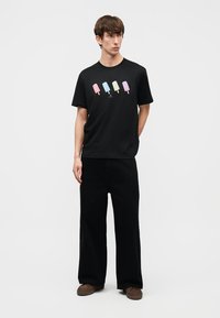 Νεαρός άνδρας φοράει μαύρο T-shirt με τέσσερα παστέλ παγωτά, μαύρο παντελόνι με φαρδιά μπατζάκια και καφέ παπούτσια, στέκεται μπροστά σε ένα απλό λευκό φόντο.