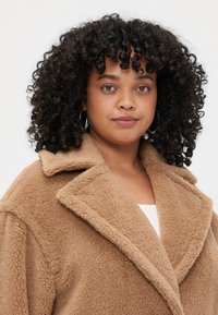 Casaco teddy oversized em tom de bege com gola com lapelas, tecido texturizado e grandes lapelas; possui uma aparência suave e peluda, adequada para o tempo frio.