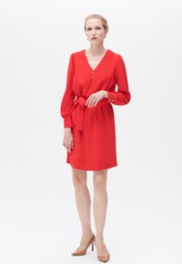 Rotes Kleid mit V-Ausschnitt, langen Ärmeln und Knopfdetails. Es verfügt über eine taillierte Taille und eine glatte Textur, kombiniert mit hellbraunen High Heels.