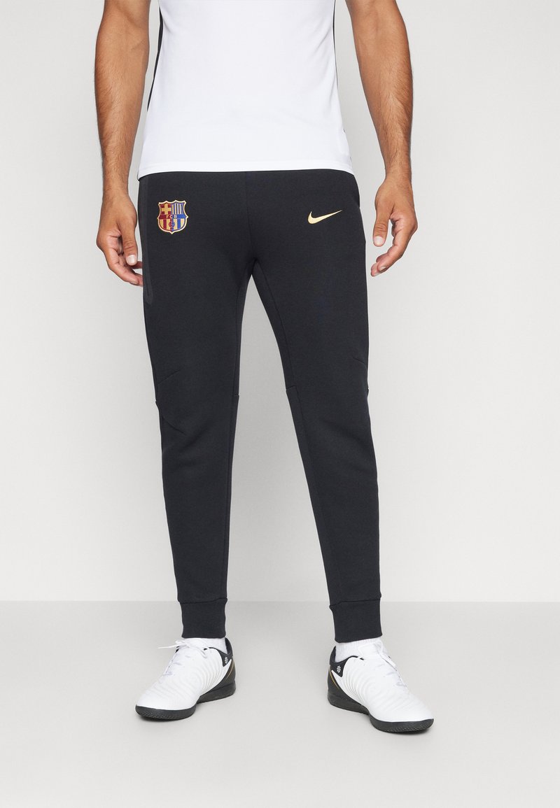 Zwarte sportbroek met een taperende pasvorm, met een gouden Nike-logo en een kleurrijk FC Barcelona-embleem op de linkerdij.