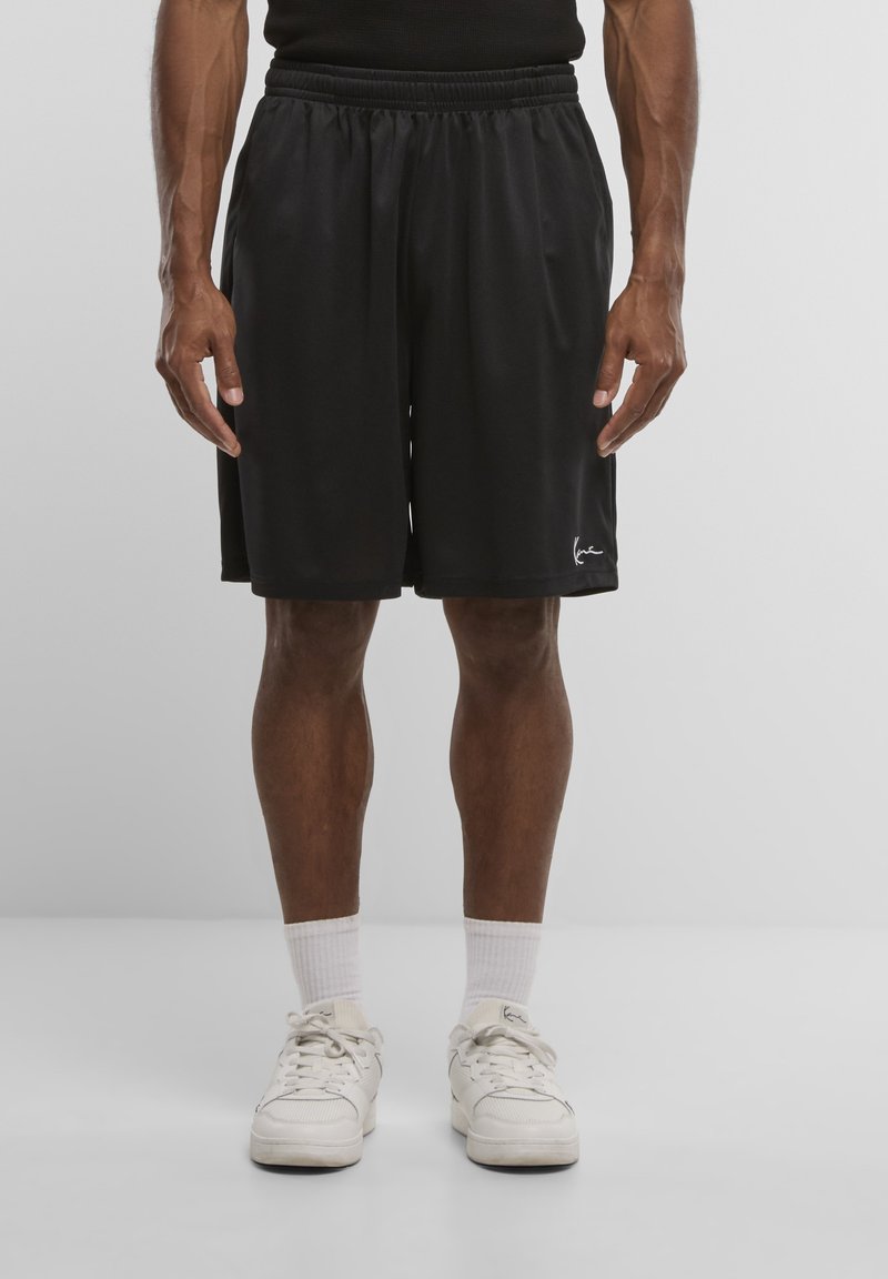 Homme portant un short de sport noir, des chaussettes blanches et des baskets blanches, debout devant un fond clair uni.