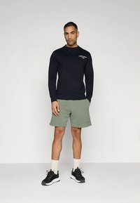 Μαύρο φούτερ με λογότυπο, πράσινο σορτς και μαύρα αθλητικά παπούτσια. Το μοντέλο στέκεται μπροστά σε απλή φόντο, παρουσιάζοντας αθλητικά ρούχα για καθημερινή χρήση.