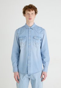 Camisa de denim azul claro con mangas largas, botones a presión, cuello puntiagudo y dos bolsillos en el pecho con detalles de costura en zigzag.