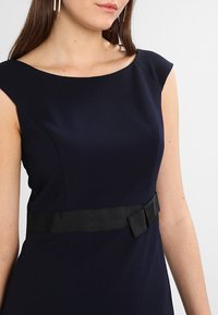 Marinblå klänning med båturringning, figurnära passform och ett svart satängbälte med en rosettaccent. Slät textur och korta ärmar.