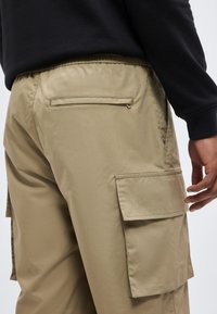 Κοντινό πλάνο ενός ατόμου που φοράει μπεζ cargo παντελόνια με πλαϊνές και πίσω τσέπες με φερμουάρ, και μαύρο μακρυμάνικο τοπ.