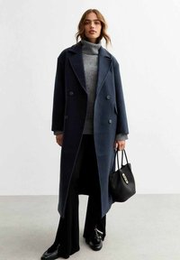 Cappotto di lana navy con chiusura doppiopetto, ampi risvolti e tasche laterali, indossato sopra un dolcevita grigio e pantaloni neri, con una borsa nera.