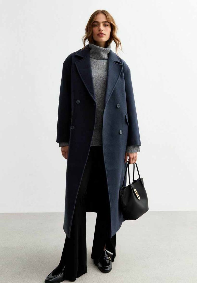 Cappotto di lana navy con chiusura doppiopetto, ampi risvolti e tasche laterali, indossato sopra un dolcevita grigio e pantaloni neri, con una borsa nera.
