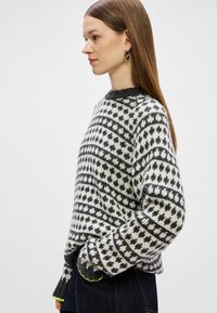 Pulover tricotat în crem și negru cu modele geometrice, având un guler ribat în gri închis și accente neon verzi la manșete.