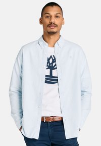 Uomo con le mani in tasca che indossa una camicia azzurro chiaro con bottoni sopra una maglietta bianca con logo di un albero blu scuro, pantaloni scuri e cintura marrone.