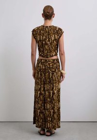 Femme avec les cheveux en chignon portant un haut sans manches à motifs bruns et une jupe longue assortie, se tenant face à un mur gris clair uni.