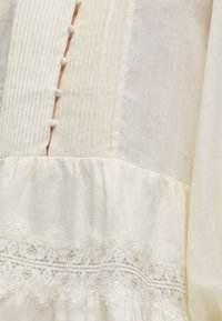 Κοντινή λήψη υφάσματος σε εκρού χρώμα με κάθετες πιέτες, κουμπιά καλυμμένα με ύφασμα, ραφή με σούρα και περίτεχνη διακόσμηση με κεντητή δαντέλα σε μορφή λουλουδιών.