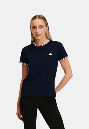 Donna che indossa una maglietta blu navy a maniche corte con bordo a righe rosse e bianche e pantaloni neri, in piedi con la mano sul fianco.
