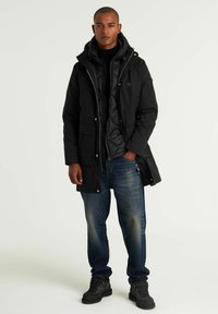 Juodas paminkštintas parka su užtrauktuku, reguliuojamu gobtuvu ir užsegamomis kišenėmis, uždėtas ant juodos laukinės liemenės. Dėvima su mėlynomis džinsinėmis kelnėmis ir juodais batais.