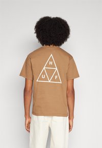Camiseta de algodón marrón con un gran logotipo triangular blanco que muestra las letras H, U y F en la parte de atrás. Mangas cortas y corte estándar.