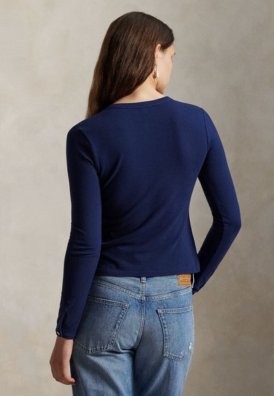 Sieviete ar gariem brūniem matiem, valkājot tumši zilu garroku augšdaļu un gaiši zilas džinsas, skatās prom pret neitrālu fonu.