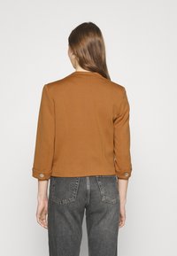 Brązowa krótka kurtka z rękawami do trzy czwarte długości, z guzikami na mankietach, zestawiona z ciemnoszarymi jeansami z wysokim stanem.