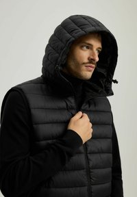 Czarna, pikowana kamizelka z kapturem, z zapięciem na zamek błyskawiczny i ściągaczami przy rękawach. Materiał jest gładki, a sekcje pikowania zapewniają izolację.