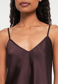 Kobieta nosząca ciemnofioletowy, satynowy top na cienkich ramiączkach z dekoltem w kształcie litery V, odsłaniający szyję, ramiona i górną część klatki piersiowej.