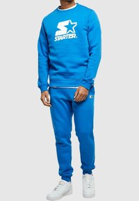 Felpa in pile blu con pantaloni abbinati, con un logo bianco. La felpa ha un collo a giro e polsini a costine; i pantaloni sono aderenti. Sneakers bianche.