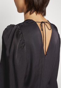 Femme portant un chemisier noir texturé avec des manches bouffantes et un dos plongeant attaché par un petit nœud en ficelle noire.
