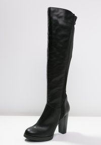 Stivale in pelle nera al ginocchio con tacco massiccio e punta tondeggiante, in posizione verticale su una superficie bianca contro uno sfondo grigio chiaro.