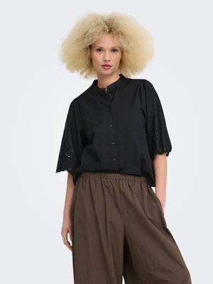 Jeune femme aux cheveux blonds bouclés porte un chemisier noir boutonné avec des manches ajourées et un pantalon marron ample taille haute, debout devant un fond blanc.