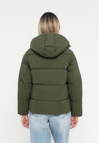 Persona con cabello ombre claro que lleva una chaqueta acolchada verde oliva con capucha, combinada con jeans descoloridos de un azul claro, de espaldas a la cámara.