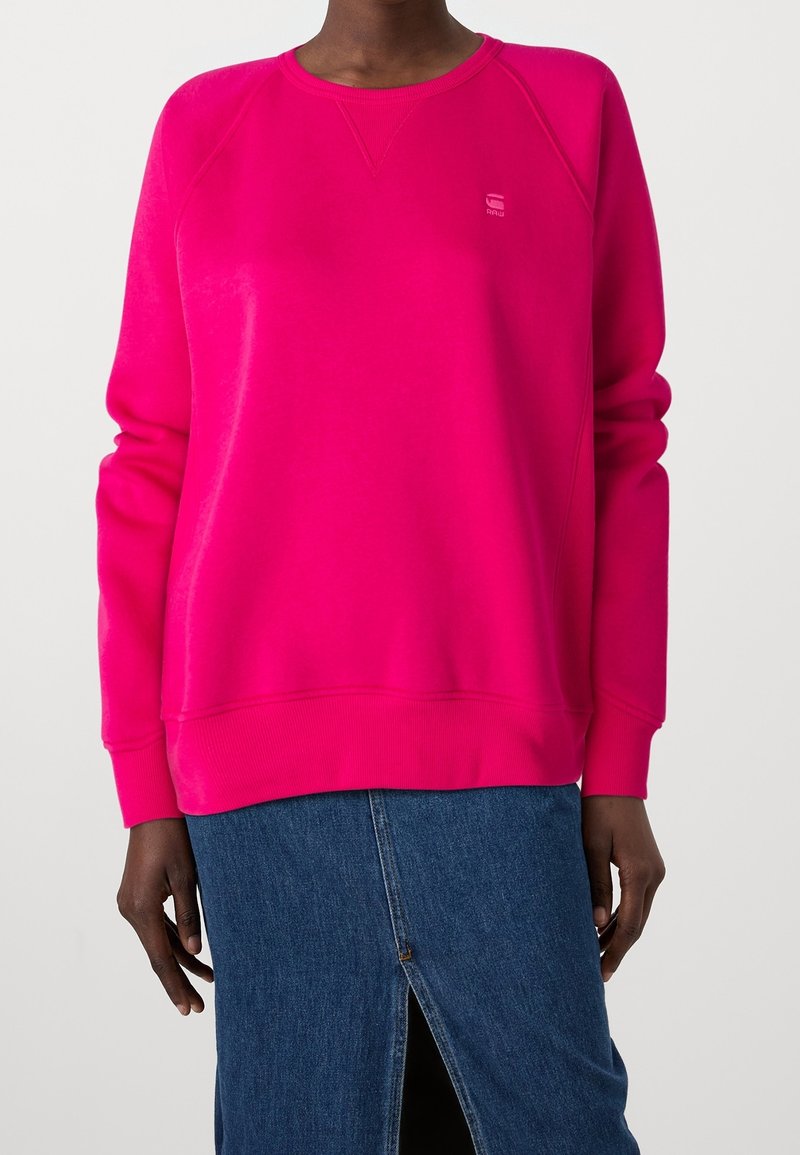 Personne portant un sweat-shirt à manches longues rose vif et une jupe en jean bleue avec une fente à l'avant sur un fond uni.