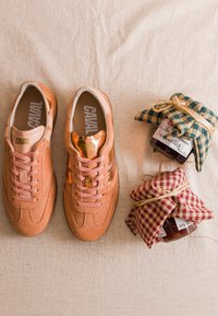 Paire de baskets CAVAL marron clair avec lacets à côté de deux pots de confiture recouverts de tissu à carreaux et liés avec de la paille sur un tissu beige.