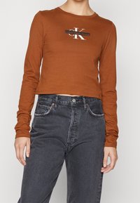 Hnědý dlouhý rukáv crop top s centrálním černobílým logem Calvin Klein, spárovaný s vysokými tmavými džíny.