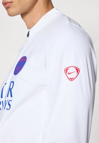 Bijela sportska majica s plavim logotipom Pariza i crvenim Nike emblema na lijevoj rukavu. Dizajn uključuje visoki ovratnik i laganu tkaninu.