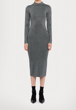 Woman wearing a fitted, long-sleeve, high-neck, ankle-length silver-gray dress with pointed black ankle boots against plain background.