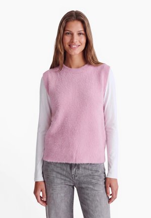 Jeune femme aux longs cheveux bruns portant un haut rose sans manches en tissu pelucheux par-dessus une chemise blanche à manches longues et un jean gris, souriante à la caméra.