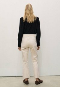 Beige hoge taille broek met een rechte snit, met een rafelige zoom, gedragen met een zwart shirt met lange mouwen en bruine schoenen, gezien van achter.