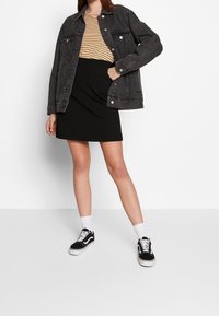 Giacca di jeans nera sopra una maglietta a righe gialle e nere, abbinata a una gonna nera e sneakers nere. Calzini bianchi visibili. Design semplice.