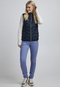 Gilet matelassé bleu marine avec motif en chevrons, fermeture éclair argentée, porté sur un haut à manches longues de couleur claire, associé à un pantalon violet et des baskets neutres.