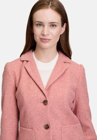 Vaaleanpunainen villatakki, jossa on kaarrelta sijaitsevat läpipainolevyt, kaksi etutaskua ja kaksi pyöreää nappia. Pehmeä rakenne ja istuva malli.