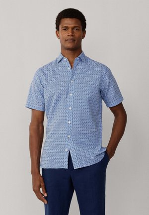 Mann trägt kurzärmeliges blau gemustertes Hemd mit Knöpfen und dunkelblaue Hose, steht mit einer Hand in der Tasche vor einfachem Hintergrund.