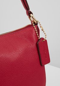 Sac à main en cuir rouge avec une finition texturée, une fermeture éclair dorée et un accent en chaîne. Comprend une étiquette avec un logo embossé et une attache de sangle robuste.