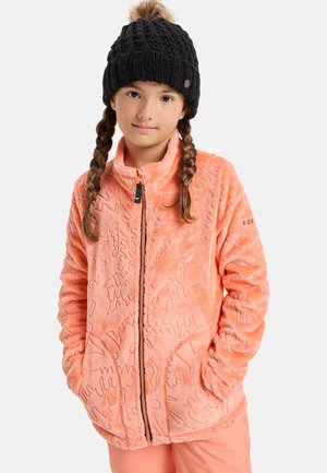 Niña con trenzas, lleva un gorro de punto negro y una chaqueta polar coral con cremallera, con las manos en los bolsillos, sobre un fondo blanco.