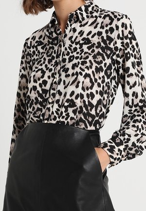 Kvinde iført en langærmet leopardmønstret skjorte med knapper, stoppet ned i en højtaljet sort lædernederdel med hånden i lommen.