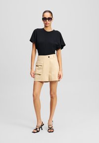 Top nero a maniche corte abbinato a una gonna cargo beige con tasca laterale e chiusura a bottone, completato da sandali neri con tacco e cinturini.