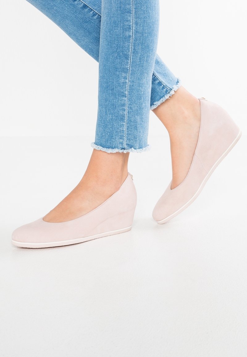 Pieds chaussés de chaussures compensées rose clair, portées avec un jean denim bleu coupé, sur un sol et un fond blancs.