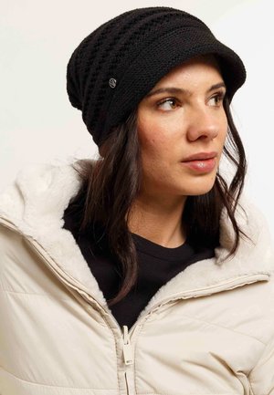 Donna con capelli scuri che indossa un berretto di maglia nero con un piccolo logo e una giacca color crema con colletto foderato di pelliccia, che guarda di lato.