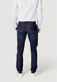 Jeans de mezclilla azul oscuro con un diseño ajustado, que cuentan con bolsillos traseros y una etiqueta de cuero marrón. Combinados con una camisa blanca de manga larga.