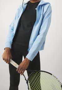 Sudadera con cremallera de color azul claro con bolsillos frontales, superpuesta a una camiseta negra. Leggings negros y una raqueta de tenis con un agarre estampado son visibles.