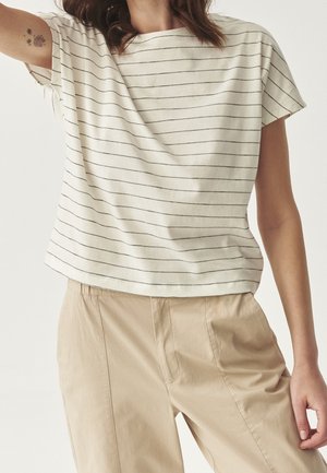 Femme portant une chemise blanche à manches courtes avec de fines rayures horizontales noires et un pantalon beige taille haute sur un fond uni.