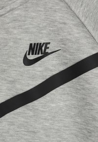 Šedá mikina s hladkým povrchom, čiernym logom Nike a diagonálnym čiernym pruhom cez oblasť hrudníka.