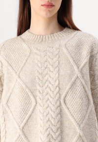 Beige neule, jossa on teksturoidut neulekuviot, sisältäen kaapeli- ja timanttimuotoja. Ribbattu pyöreä kaula-aukko. Pehmeä, paksu materiaali.